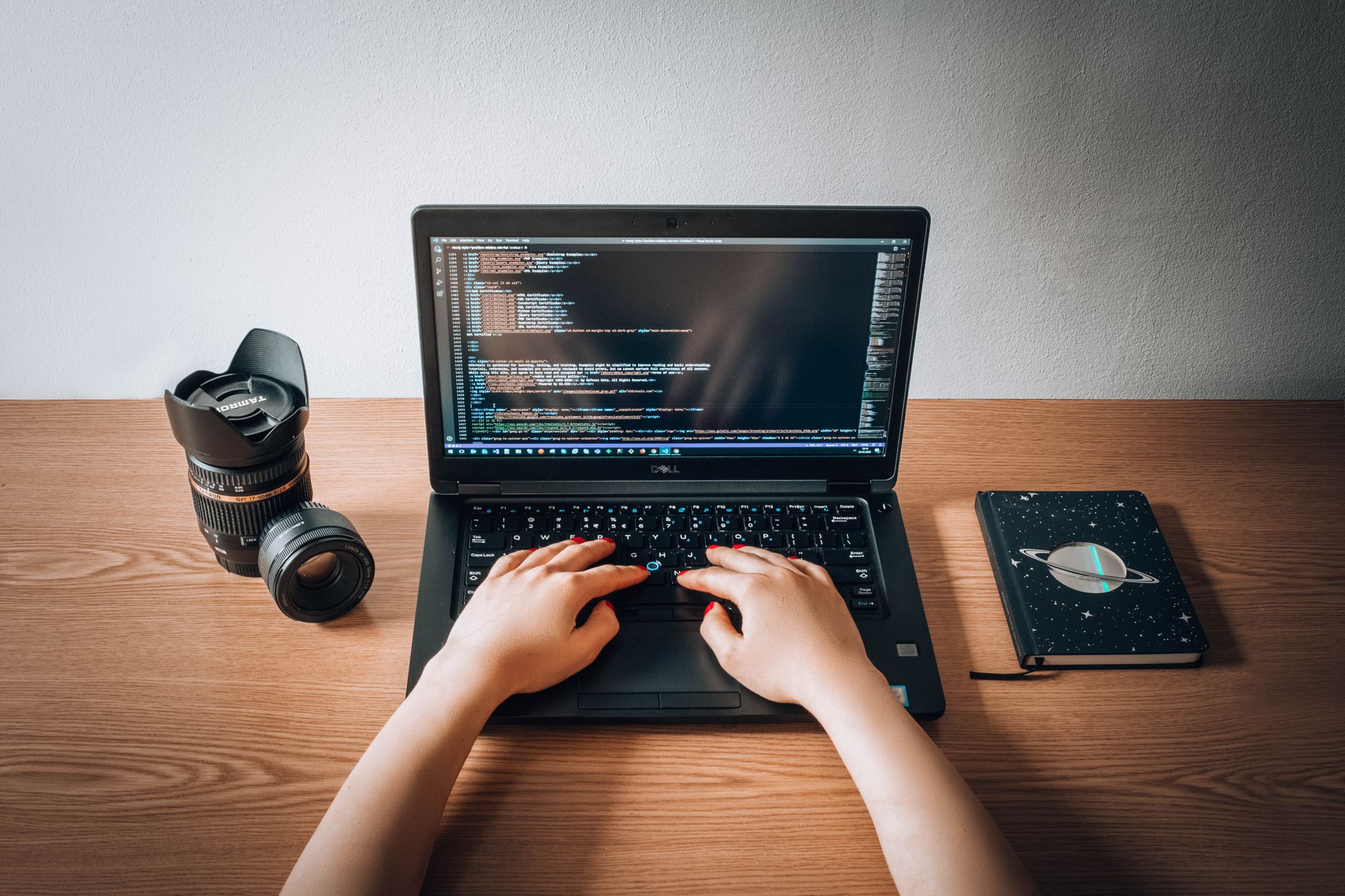 A person works on a laptop at a wooden desk with a notebook and camera lens.